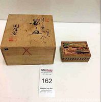 Two wooden boxes placed side by side on a white surface; the larger is plain wood with writing, the smaller decorated with intricate colorful folk art