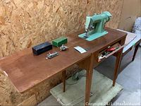 Full side-angle view of vintage Singer sewing machine mounted in wooden cabinet table with flat sides extended, showing sewing attachments on top and drawer open with tools.