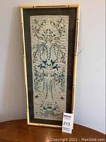 Photo showing full view of the framed silk embroidery textile art standing on a wooden surface against a white wall.