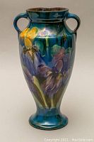 Front view of blue glazed Torquay pottery urn with dual handles and floral motif in yellow and purple.