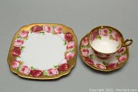 Square luncheon plate with rose and gold trim alongside a teacup and matching saucer all set against a neutral background.
