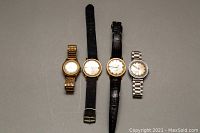 Four vintage men's wristwatches displayed horizontally: two with black leather straps, one with gold-tone expandable metal bracelet, and one silver-tone metal link bracelet. Various dial and case designs.