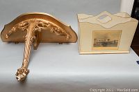Side view of ornate gold wall shelf showing carved decorative design and magazine rack with scenic panel on side.