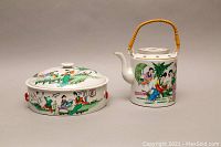 Front view of hand painted Asian porcelain kettle with bamboo handle and matching covered dish, both decorated with traditional figures and nature scenes.