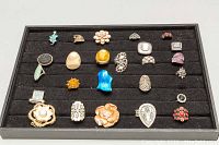 Full view of 25 assorted costume rings organized in a black felt tray, showing various designs, colors, and styles including floral, geometric, and stone-set rings.