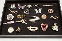 Photo showing all 18 assorted brooches laid out in black case, varied designs including animal shapes, flowers, hearts, and abstract sculptures. Visible missing stones and wear present.