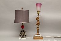 Photo showing two decorative table lamps with cherub figures and marble bases. One has a brown fabric shade and red glass accent, the other has a frosted pink glass shade with flea chips.