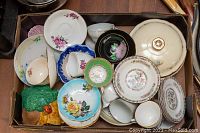 Top-down view of assorted chinaware including plates, bowls, teacups, and a lidded dish showing floral and gold accents with some damage visible.