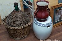 Full view of the wicker-covered glass wine carboy, ceramic plant stand with blue pattern, and reddish-brown ceramic urn grouped together on wooden floor.
