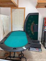 Full view of oval foldable poker table with green felt and separate blackjack table top leaning against wall.