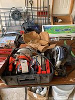 Wide view of black plastic carrying case containing Black & Decker plunge router, leather work belt, circular saw, and assorted items on garage table.