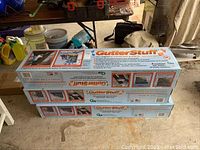 Three unopened GutterStuff boxes stacked under a workbench in a garage.