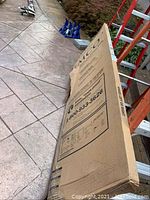 Long cardboard box labeled EMCO on tiled patio floor with ladder and jack stands in background