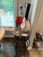 Full view of granite top plant stand with red glass, abstract white and black vase, and decorative ceramic vase with blue floral design in background.