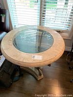 Full view of one round end table showing beige marble top with glass center insert, on wood floor near window with blinds.