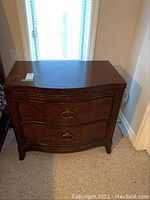 Front view of one wooden chest-style nightstand showing curved front and two drawers with metal ring pull handles