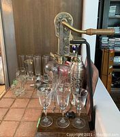 Photo showing vintage metal bottle press, several stemmed glasses, tall glasses, and metal cocktail shaker on tile countertop.