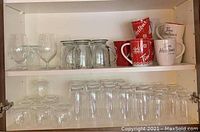 Two‐shelf view showing top shelf stemmed glasses, mason‐style mugs, Tim Hortons mugs; bottom shelf inverted tumblers