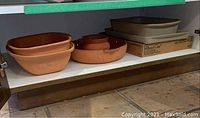 Shelf view showing two grey Pampered Chef stoneware pans on right and three terracotta clay dishes on left