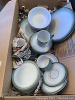 Photo of boxed set of Denby stoneware dishes including multiple plates, bowls, cups, and saucers with visible wear and newspapers used as packing material.