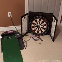 Shows the dart board cabinet with doors open, dart board centered, and two sets of darts on the floor. The electronic putting game is placed beside the dart board on the carpeted floor with visible digital display and putting green mat.