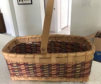 Image of a hand-woven rectangular market basket with natural and reddish-brown materials and a single wooden swing handle.