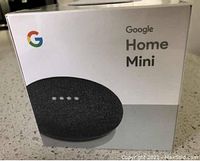 Photo showing the Google Home Mini speaker in its original box with Google branding and image of the charcoal fabric speaker.