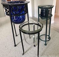 Photo showing three wrought-iron candle holders with glass inserts placed on a countertop. Two have clear glass inserts; one has a blue mottled glass insert. The holders have tripod legs with decorative iron bands.