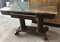 Front side view of the antique library table showing the glass top, narrow drawer, and two cylindrical pedestal legs on a carved base.
