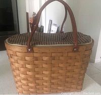 Large Longaberger Boardwalk basket with brown leather handles, checkered zippered liner, and protective cover shown from front angle on white countertop.