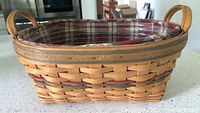 Photo of a woven Longaberger basket with oval shape, solid wooden handles, red plaid liner and clear plastic protector inside, showing condition and liner details