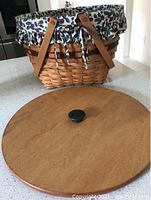 Light brown woven wooden basket with two tan leather handles on each side. Inside the basket is a white fabric liner with a holly berry print, partially visible tied to the basket. In front of the basket is a smooth round wooden lid with a black knob handle attached.