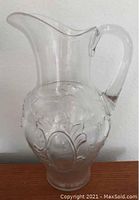 Clear glass pitcher with ornate embossed patterns and handle, standing approximately 12 inches tall.