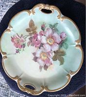 Top side of a decorative painted china plate with floral artwork in pink and white flowers with green leaves and scalloped gold-edged rim