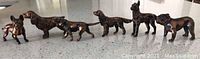 Six heavy metal dog figurines lined up on a floor surface, showing different dog breeds and poses, metallic brown finish, approx. 2-3 inches each.