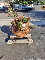 Front view of round terracotta pot on iron stand with red-flowered plant inside, showing clay flaking and rusted iron.