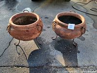 Two terracotta pots with iron ring handles and stands on pavement showing overall condition and size.