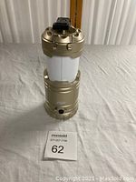 Front view of a gold Flipo Slide-N-Go LED lantern standing upright on a table with a MaxSold label 62 in front.