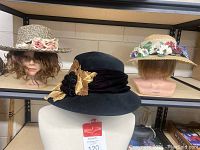 Three vintage hats displayed on mannequin heads: black Bailey Tomlinson hat with gold decorations, woven Talbots hat with roses, straw hat with colorful flowers on rim.