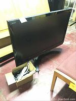 Front angle showing the screen, base stand, and a small box with cables on the floor next to the TV.