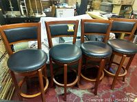 Four solid brown wood swivel barstools with black textured padded seats and backs, brass tack detail around seat edges.