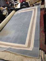 Large rectangular rug with solid blue center and white and tan border laid out partially unrolled indoors on a floor.