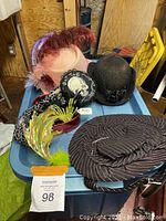 Photo showing six assorted women's costume hats placed on a blue surface, including pink felt hat with feathers, black lace hat with cameo, burgundy velvet hat with green feather decoration, and a black pinstriped fabric hat.