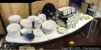 Overview of the set showing stacks of white plates, cups, mugs, and glassware on the table.