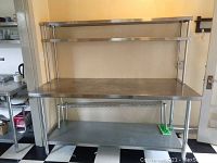 Stainless steel prep tables next to wall showing dual-tier overhead shelves and bottom panels with wire shelf on lower rack