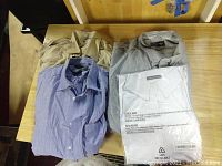 Four men's dress shirts displayed on a wooden table showing colors and condition. Beige, light blue, gray, and white color shirts, with the white shirt in packaging.
