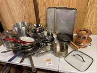 Full assortment of pots, pans, baking trays, copper gadget, and waffle iron arranged on table.