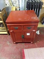Front view of red wooden side table with two drawers and double-door cabinet below on a concrete floor with some adjacent furniture and folded chairs in background.