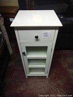 Front view of small white cabinet showing drawer and glass door with two shelves inside.