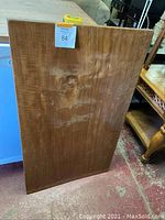 Solid rectangular wood tabletop standing upright showing brown wood surface with visible wood grain and knots, and scratches on the surface.